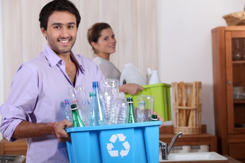 A young man with dark hair and a light purple shirt, smiling at the camera while holding a blue recycling bin filled with empty glass bottles and jars, situated in a domestic kitchen with a woman in the background, who is looking to the side and appears to be sorting or preparing clutter. The background includes wooden kitchen furniture, such as a glass-fronted cabinet and a basket, with a light-colored curtain or wall. The scene depicts a clear focus on recyclable waste collection within a home environment, consistent with rubbish removal services offered by Office Clearance Merton in the Merton area, with natural lighting highlighting the textures of the glass bottles and the smooth finish of the bin.