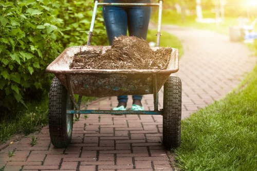A person wearing blue jeans and dark footwear pushing a rusted metal wheelbarrow filled with dark, moist soil along a paved pathway bordered by green bushes on the left and a grassy area on the right. The wheelbarrow's handles are metallic with plastic grips, and the large black rubber tire is centered beneath the metal basin. In the background, the pathway extends into a residential area with a parked car visible further along, suggesting a suburban environment possibly within the Merton postcode area. The scene is illuminated by natural daylight, highlighting the earthy textures of the soil and the weathered surface of the wheelbarrow, aligning with typical rubbish and waste removal activities that Office Clearance Merton offers for garden and outdoor clearing jobs in the local community.