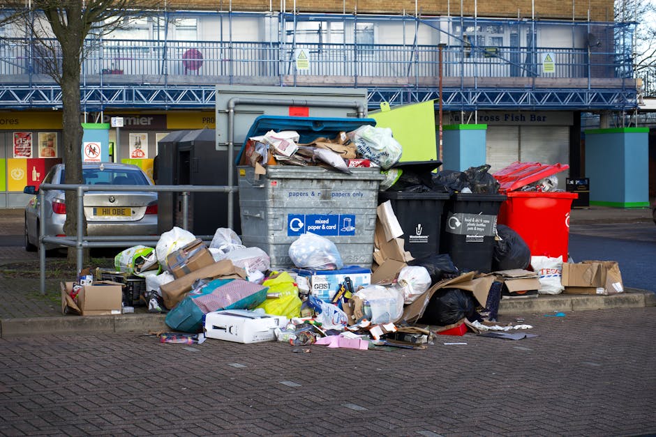 A pile of black garbage bags, cardboard boxes, and discarded packaging materials stacked outside a building against a textured, off-white wall. The bags appear full and some are tied at the top, with