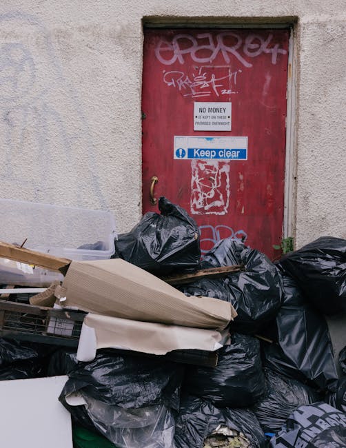 A pile of black rubbish bags and discarded debris stacked against a textured concrete wall with a red metal door behind, which is covered in graffiti and has a white sign indicating no money exchange 