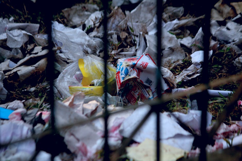 A pile of black rubbish bags and discarded debris stacked against a textured concrete wall with a red metal door behind, which is covered in graffiti and has a white sign indicating no money exchange 